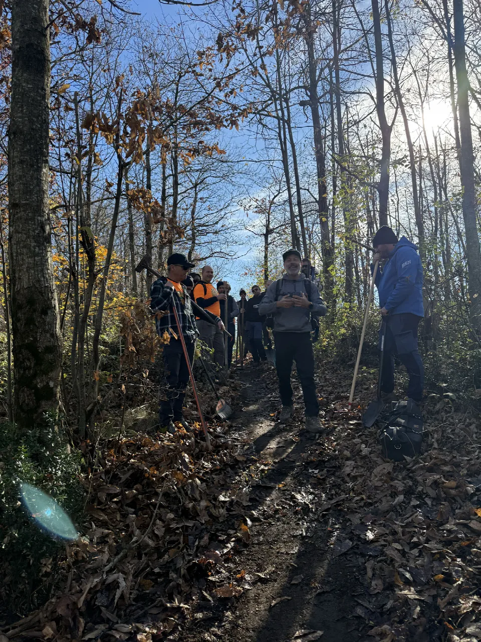 Un gruppo di persone su un sentiero nel bosco, con foglie secche a terra e alcuni con pale in mano